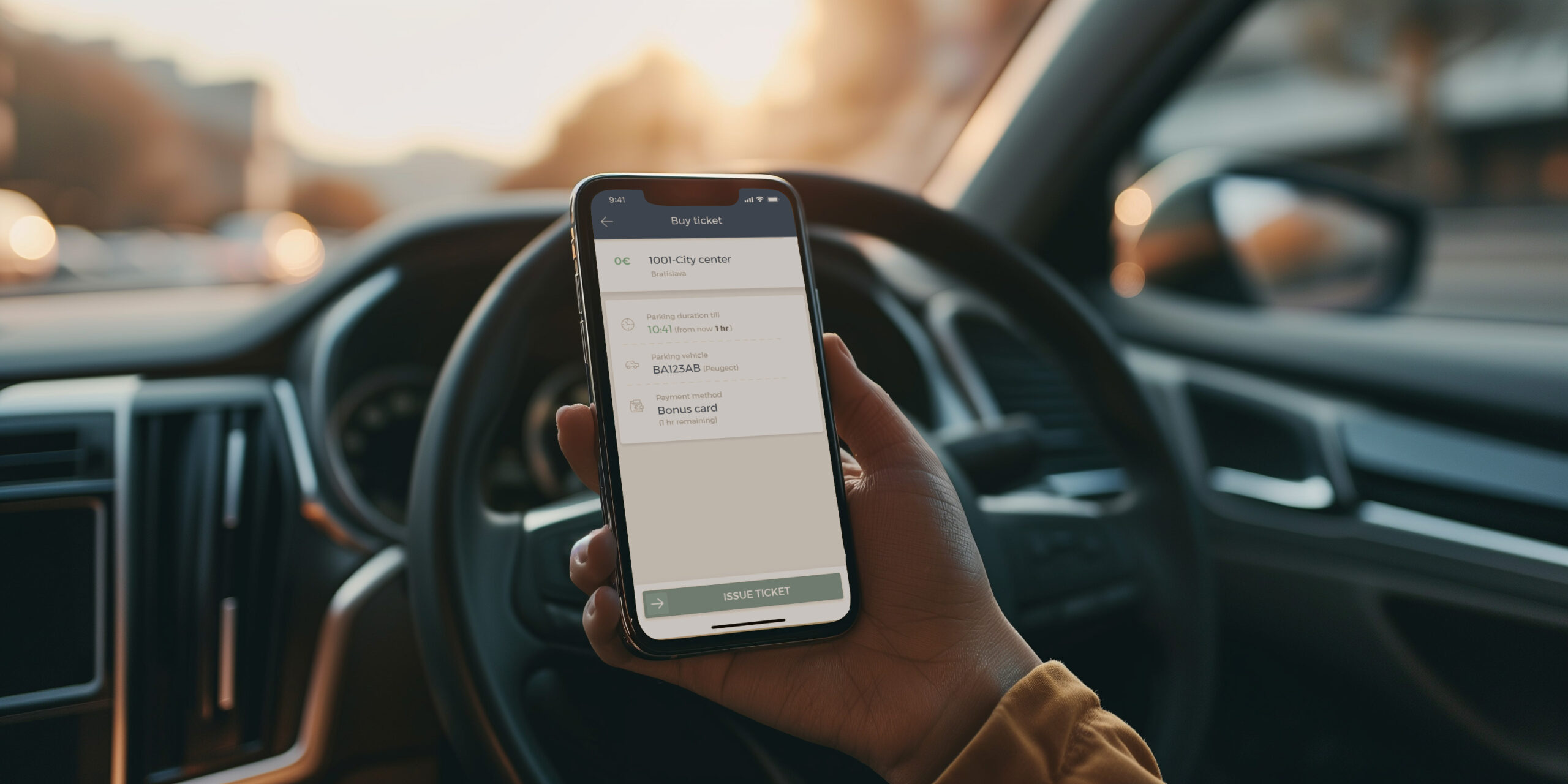 Close-up of a person's hand holding a smartphone with a parking ticket app open, inside a car. The screen shows options for parking duration and vehicle information against a blurred backdrop of a car's interior and a city street at sunset.