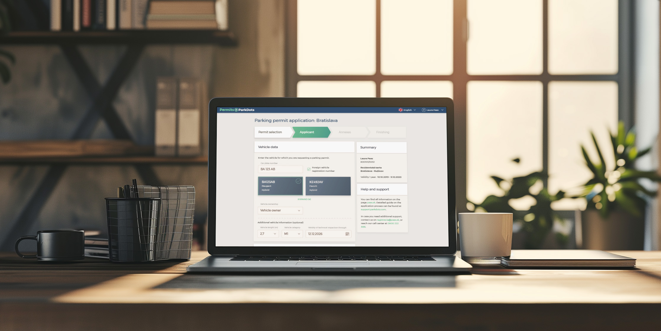 A laptop on a wooden desk displaying a parking permit application screen, with sections for vehicle data and summary visible. The desk is set against a backdrop of a bright window and bookshelf, with a mug and notepad alongside the laptop.